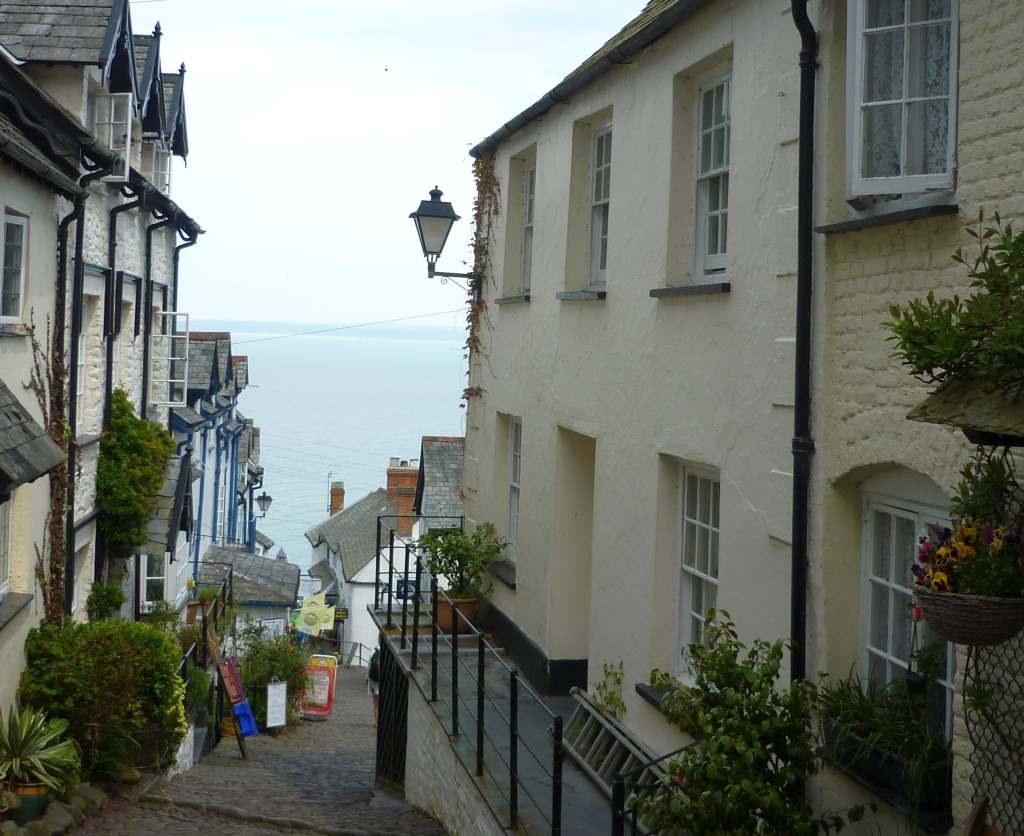 A steep narrow lane between white/cream cottages and shops to the sea beyond.