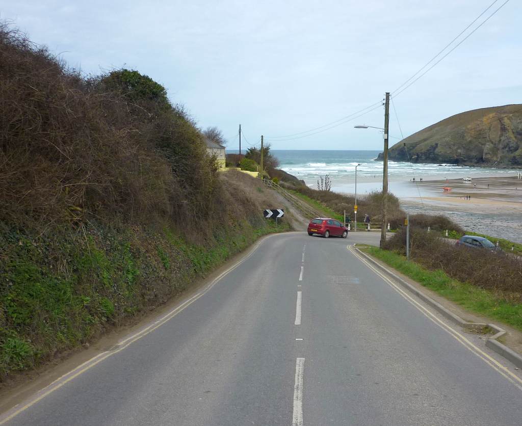 Looking down a narrow road that meets a sandy beach, the sea and a grassed cliff beyond.