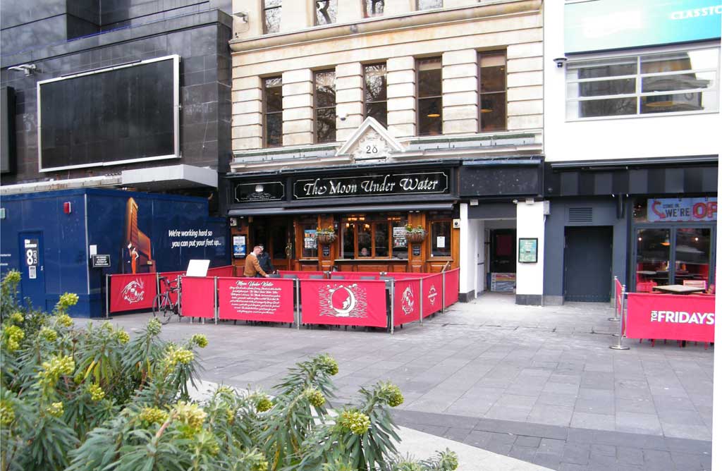 A row of shops in Leicester Square the main one being "The Moon Under Water" with plants in the foreground