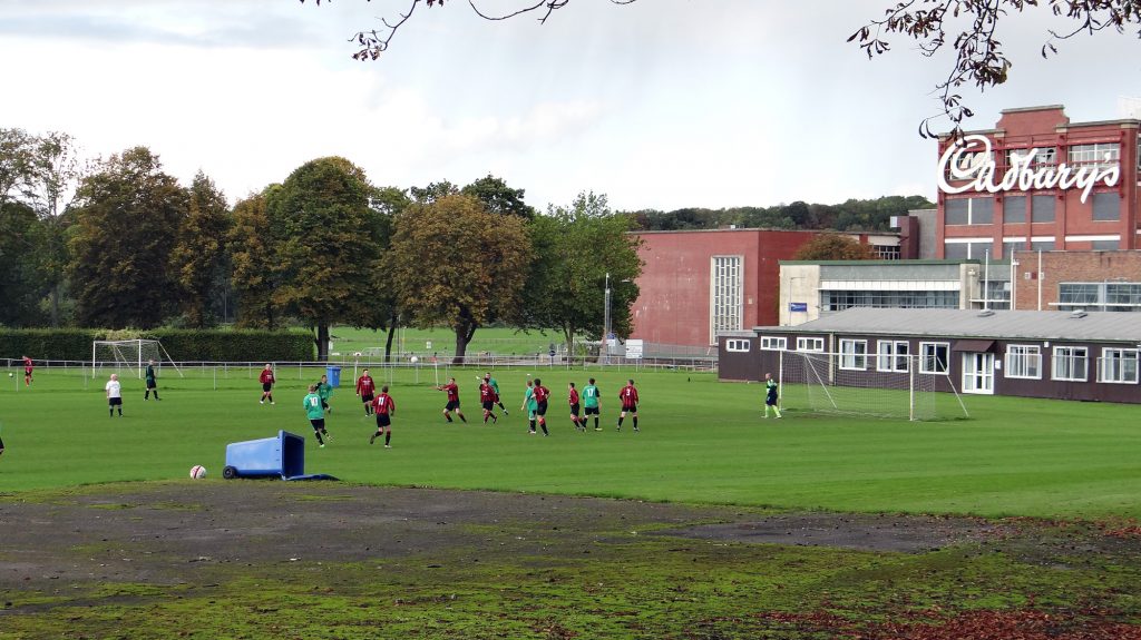 A football field with men playing against the background of a club house and factory building with the name Cadbury's emblazoned on it