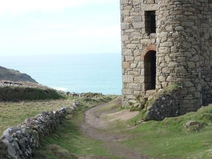 A view across a cliff top to the sea with a well maintained stone building to the right being the remains of a Cornish mine engine house.