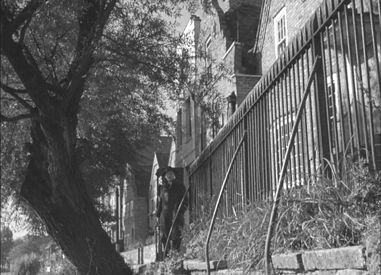 A man looking towards camera by some railing by the bank of the River Thames with houses backing him.