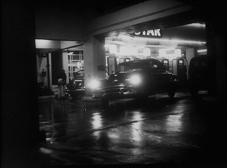 An American car pulling away from a garage with petrol pumps at night.