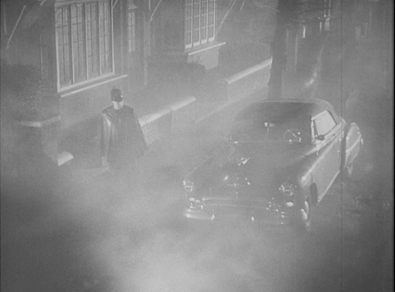 A misty picture of a police constable with a cape passing an American car by the side of the road.