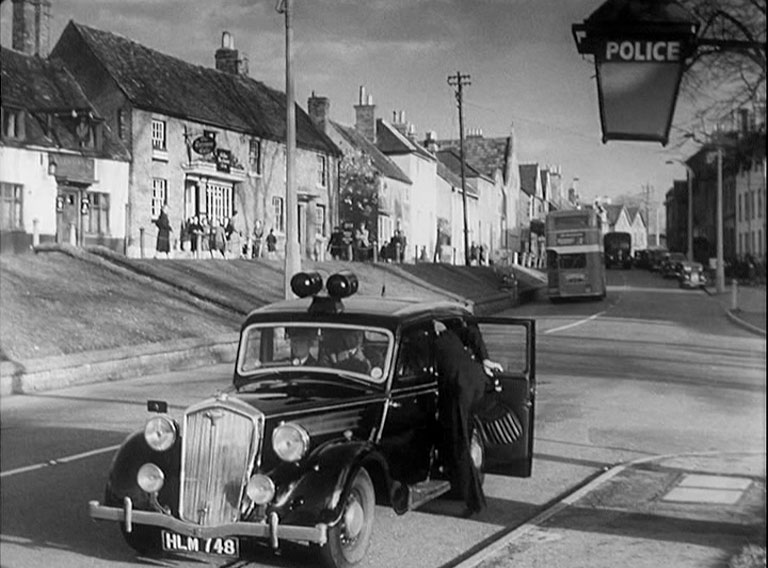 A figure entering a police car by the side of a road against the view of a country town high street. The Shute, Wokingham, looking across to The Terrace Circa 1953