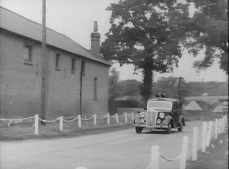 A police car approaching camera up a country lane or drive beside a farm building wall.