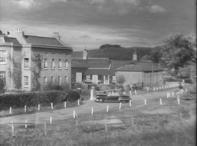 An American car pulling into the drive of a very large mansion with stables to the rear.