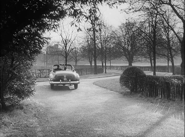 A man waving from an open top American car as it pulls away towards a tree lined drive in winter.