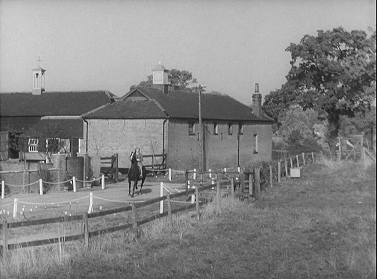 A woman on a horse coming out of a stable complex towards camera.