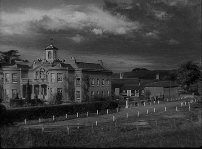 A darkened shot of a mansion with stabling to the rear.